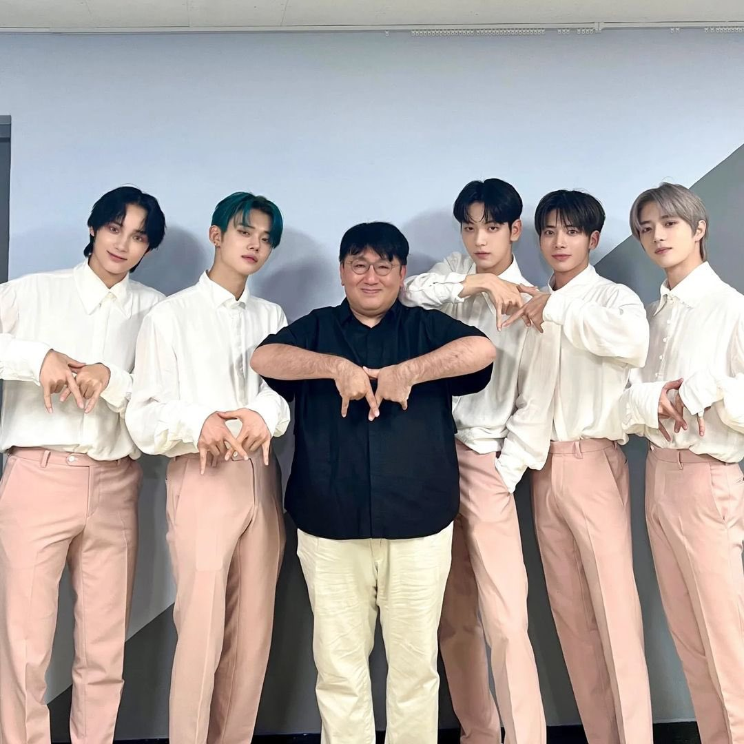 HYBE Chairman Si-hyuk Bang standing in the middle of all five members of TXT, doing their signature group hand pose